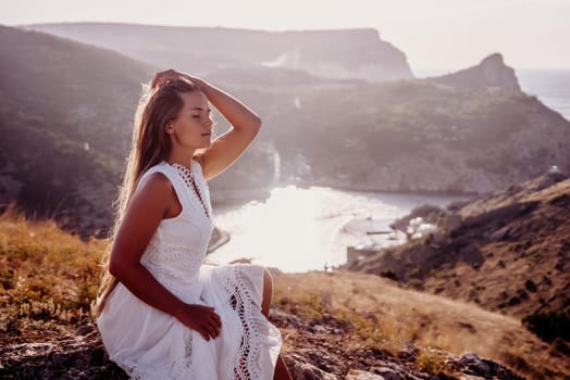 A woman in a white dress is sitting on a rock overlooking a body of water. She is enjoying the view and taking in the scenery