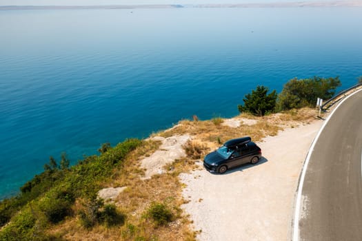 Car with a roof box, parked near a scenic cliffside road, overlooking a vast and azure sea.
