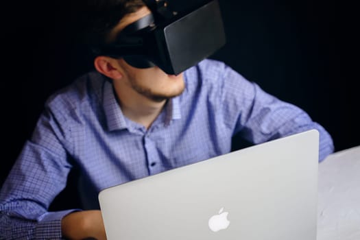 A man is wearing a virtual reality headset while seated in front of an Apple laptop, engaging in a digital experience. Bishkek, Kyrgyzstan - March 14, 2020