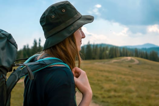 A young girl in a hat and with a backpack goes on an adventure, travels and enjoys nature, a woman looks at a picturesque view with meadows, forest and mountains on a sunny day.