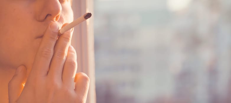 A young man smokes, holds a cigarette in his hand and inhales the smoke while standing at the window early in the morning in the sun, close-up view.