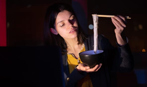 Young girl works at night at home with laptop and eats noodles, asian food with chopsticks. A woman watches a show, a movie and has dinner with ordered food from a cafe.