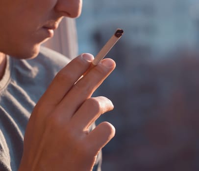 A young man smokes, holds a cigarette in his hand and inhales the smoke while standing at the window early in the morning in the sun, close-up view.