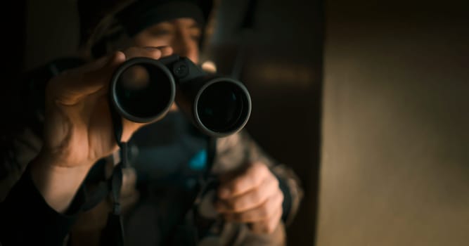 Young man in military uniform and wearing a helmet looks through binoculars during the fighting, undergoes military exercises. The soldier conducts reconnaissance, near the window inside the building.
