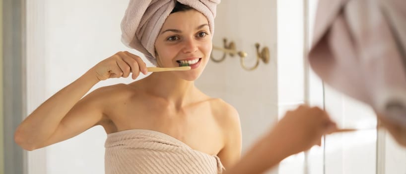 A young girl brushes her teeth near the mirror after relaxing in the bathroom, the woman is wrapped in a towel and takes care of the health and beauty of her body and teeth, gums.