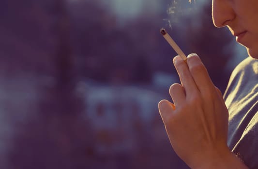 A young man smokes, holds a cigarette in his hand and inhales the smoke while standing at the window early in the morning in the sun, close-up view.