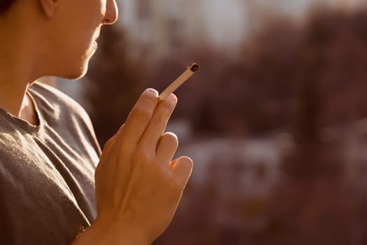 A young man smokes, holds a cigarette in his hand and inhales the smoke while standing at the window early in the morning in the sun, close-up view.