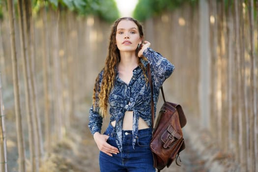 A confident young woman in a stylish denim outfit holds a chic backpack, surrounded by serene nature. Her bright smile and relaxed demeanor reflect a carefree summer lifestyle, embodying adventure