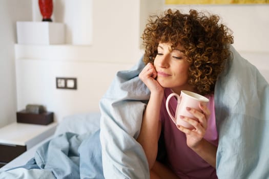 Woman wrapped in blanket enjoying hot beverage and leaning on hand while resting on bed in weekend