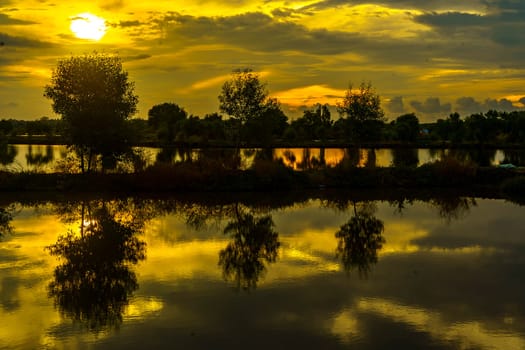 A breathtaking sunset casts golden hues over a tranquil pond near Mengare, Gresik, Indonesia. Silhouetted trees and clouds create a dramatic reflection on the water's surface.