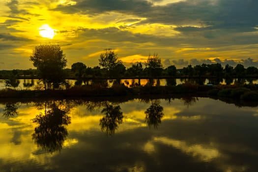 A breathtaking sunset casts golden hues over a tranquil pond near Mengare, Gresik, Indonesia. Silhouetted trees and clouds create a dramatic reflection on the water's surface.