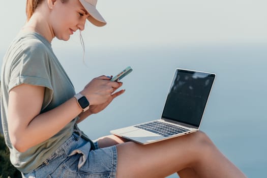 A young woman wearing a baseball cap sits outdoors, checking her phone while a laptop sits on her legs.