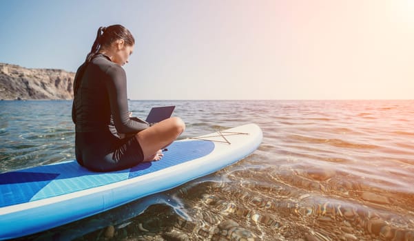 Digital nomad, woman in the hat, a business woman with a laptop sits on the rocks by the sea during sunset, makes a business transaction online from a distance. Freelance, remote work on vacation.