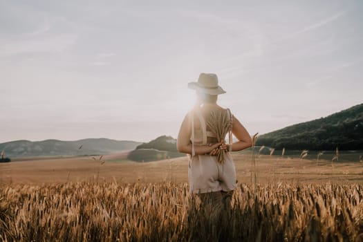 A woman wearing a hat and a light-colored dress stands in a field of tall golden wheat. The sun shines brightly behind her, casting a warm glow on the scene.