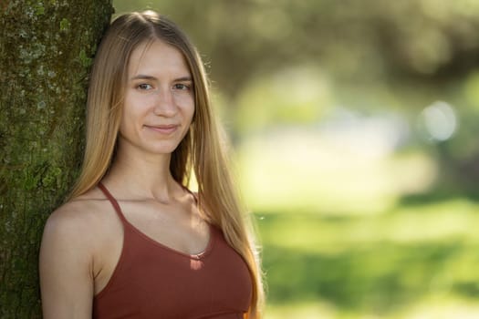 Young woman with long blond hair wearing sportswear, smiling and leaning against a tree in a park