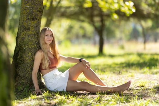 Young woman is sitting under the shade of a tree in a park, enjoying the summer weather