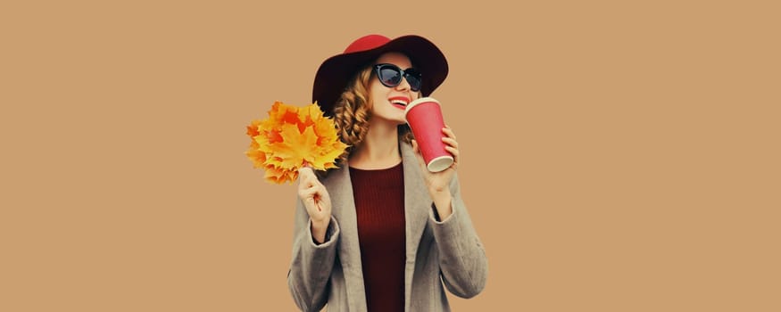 Autumn portrait of beautiful smiling young woman with yellow maple leaves, cup of coffee drink, stylish girl in hat looking away on brown background