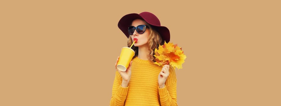 Autumn portrait of beautiful young woman with yellow maple leaves, enjoying cup of coffee drink, stylish girl in hat posing on brown background