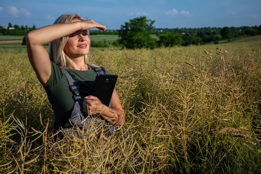 In the middle of the rapeseed field, a woman farmer stands, holding a tablet close to her body as she checks the quality of the crop. With one hand on the tablet, she gazes into the distance, her eyes fixed on the expansive yellow landscape stretching out before her. Experienced and dedicated, she takes pride in caring for her crops in the vast field. Her blonde hair catches the sunlight as she smiles with satisfaction, though the sun's rays also glare down upon her, causing her to squint slightly and shield her eyes with her hand. Her white teeth are visible. The fertile soil has proven productive once again, yielding yellow rapeseed ready for harvest. With her tablet in hand and a sense of fulfillment in her heart, the woman farmer embraces the bountiful and lavish harvest. Clad in working clothes, her sun-kissed skin radiates contentment as she stands amidst the fields. Under the blue sky adorned with fluffy white clouds, her spirit merges with the warmth of the sun, celebrating the culmination of her dedication and the richness of the land's yield