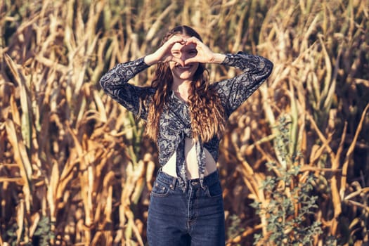 In a beautiful and vibrant golden field, a lively young woman effortlessly forms a heart shape using her hands, which wonderfully captures her infectious happiness and profound sense of contentment
