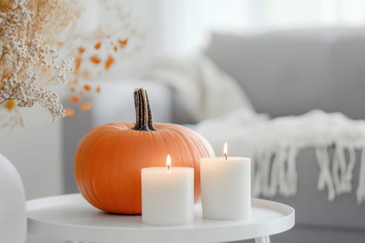 A warm atmosphere fills the living room where a small pumpkin rests next to a lit candle and a vase of dried flowers on a white table, evoking the spirit of autumn.