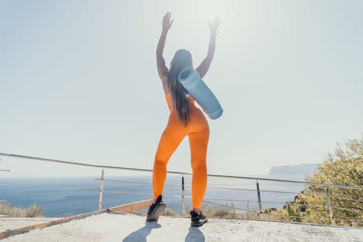 A young woman in orange yoga clothes with a blue mat slung over her shoulder raises her arms in triumph against a bright blue sky.