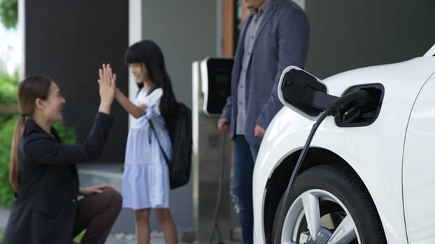 Progressive parents with electric vehicle and home charging station. Happy family with daughter giving each other high fives before leave for school. Alternative future transportation concept of EVs