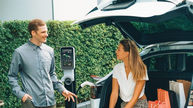 Young couple travel with EV electric car charging in green sustainable city outdoor garden in summer shows urban sustainability lifestyle by green clean rechargeable energy of electric vehicle innards