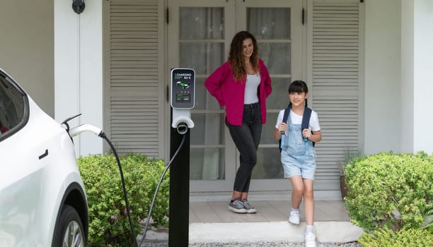 Happy little young girl learn about eco-friendly and energy sustainability as she help her mother recharge electric vehicle from home EV charging station. EV car and modern family. Panorama Synchronos