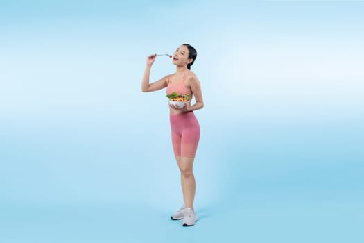 Young sporty Asian woman in sportswear holding salad bowl fill with vibrant of fruit and vegetable. Natural youthful and fit body lifestyle with balance nutrition on isolated background. Vigorous