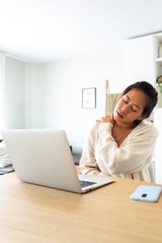 Asian woman suffering from neck pain. Chinese woman working with laptop at home self massaging neck. Vertical image.