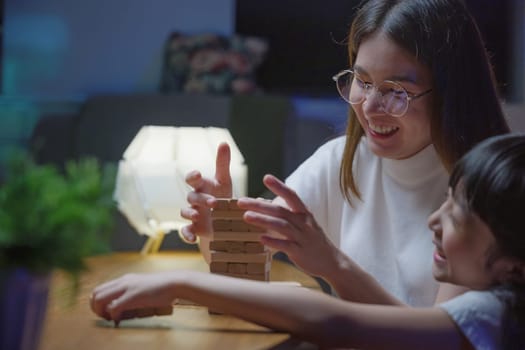 Smiling woman help teach child play build constructor of wooden blocks, Asian young mother playing game in wood block with her little daughter in home living room at night time before going to bed