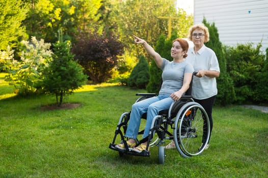 An elderly woman is carrying an adult daughter sitting in a wheelchair. Caucasian woman pointing her finger admiringly while walking outdoors