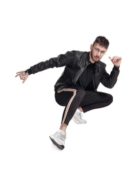 Full-length photo of a modern performer in glasses, black leather jacket, t-shirt, sports pants and light sneakers fooling around in studio. Indoor photo of a good-looking guy posing sitting and looking at the camera isolated on white background. Music and imagination.