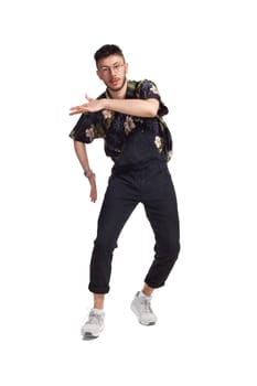 Full-length photo of an unprofessional dancer in glasses, black jumpsuit, funny dark t-shirt and sneakers fooling around in studio. Indoor photo of a guy dancing isolated on white background. Music and imagination.