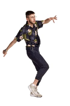 Full-length portrait of a graceful guy in glasses, black jumpsuit, colorful t-shirt and gray sneakers fooling around in studio. Indoor photo of a man dancing sideways isolated on white background. Music and imagination.