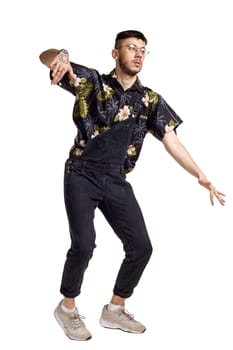 Full-length portrait of an athletic dancer in glasses, black jumpsuit, colorful t-shirt and gray sneakers fooling around in studio. Indoor photo of a man dancing isolated on white background and looking away. Music and imagination.