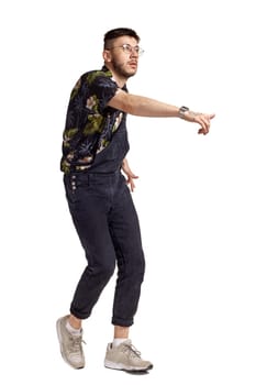 Full-length portrait of an athletic male in glasses, black jumpsuit, colorful t-shirt and gray sneakers fooling around in studio. Indoor photo of a man dancing isolated on white background. Music and imagination. Copy space.