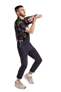 Full-length portrait of an athletic guy in glasses, black jumpsuit, colorful t-shirt and gray sneakers fooling around in studio. Indoor photo of a man dancing sideways isolated on white background. Music and imagination.