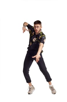 Full-length portrait of a handsome man in glasses, black jumpsuit, colorful t-shirt and gray sneakers fooling around in studio. Indoor photo of a man dancing isolated on white background. Music and imagination. Copy space.