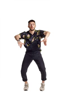 Full-length portrait of a handsome guy in glasses, black jumpsuit, colorful t-shirt and gray sneakers fooling around in studio. Indoor photo of a man dancing isolated on white background. Music and imagination.