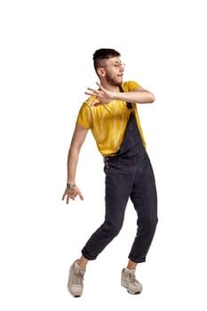 Full-length portrait of a sporty dancer in glasses, black jumpsuit, yellow t-shirt and gray sneakers fooling around in studio. Indoor photo of a man making dancing elements isolated on white background and looking away. Music and imagination concept.