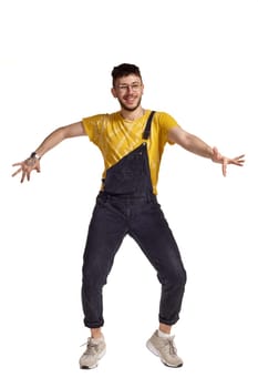 Full-length portrait of a funny man in glasses, black jumpsuit, yellow t-shirt and gray sneakers fooling around in studio. Indoor photo of a man making dancing elements isolated on white background. Music and imagination.