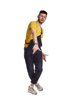 Full-length portrait of an elegant person in glasses, black jumpsuit, yellow t-shirt and gray sneakers fooling around in studio. Indoor photo of a man making dancing elements isolated on white background and looking at the camera. Music and imagination.