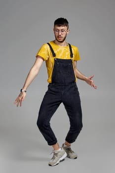 Full-length portrait of a stylish male in glasses, black jumpsuit, yellow t-shirt and gray sneakers fooling around in studio. Indoor photo of a man dancing on a gray background. Music and imagination.