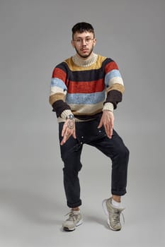 Full-length portrait of an athletic male in glasses, black jumpsuit, multi-colored sweater and gray sneakers fooling around in studio. Indoor photo of a man dancing on a gray background. Music and imagination.