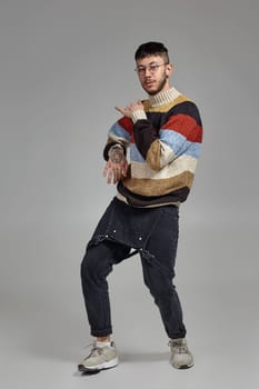 Full-length portrait of a good-looking man in glasses, black jumpsuit, multi-colored sweater and gray sneakers fooling around in studio. Indoor photo of a man dancing on a gray background. Music and imagination.
