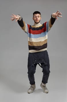 Full-length portrait of an athletic guy in glasses, black jumpsuit, multi-colored sweater and gray sneakers fooling around in studio. Indoor photo of a man dancing raising his hands on a gray background. Music and imagination.