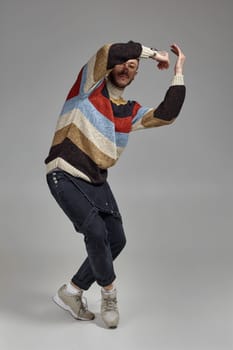 Full-length portrait of a good-looking male in glasses, black jumpsuit, multi-colored sweater and gray sneakers fooling around in studio. Indoor photo of a man dancing raising his hands on a gray background. Music and imagination.