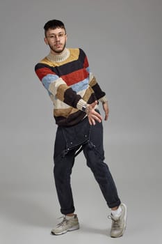 Full-length portrait of a handsome person in glasses, black jumpsuit, multi-colored sweater and gray sneakers fooling around in studio. Indoor photo of a male dancing on a gray background. Music and imagination.